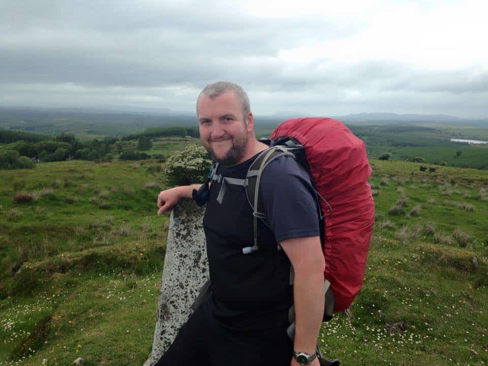 The Long Walk North – June Bright-eyed hiker in outdoor countryside with a large backpack on overcast day, standing near a stone marker.