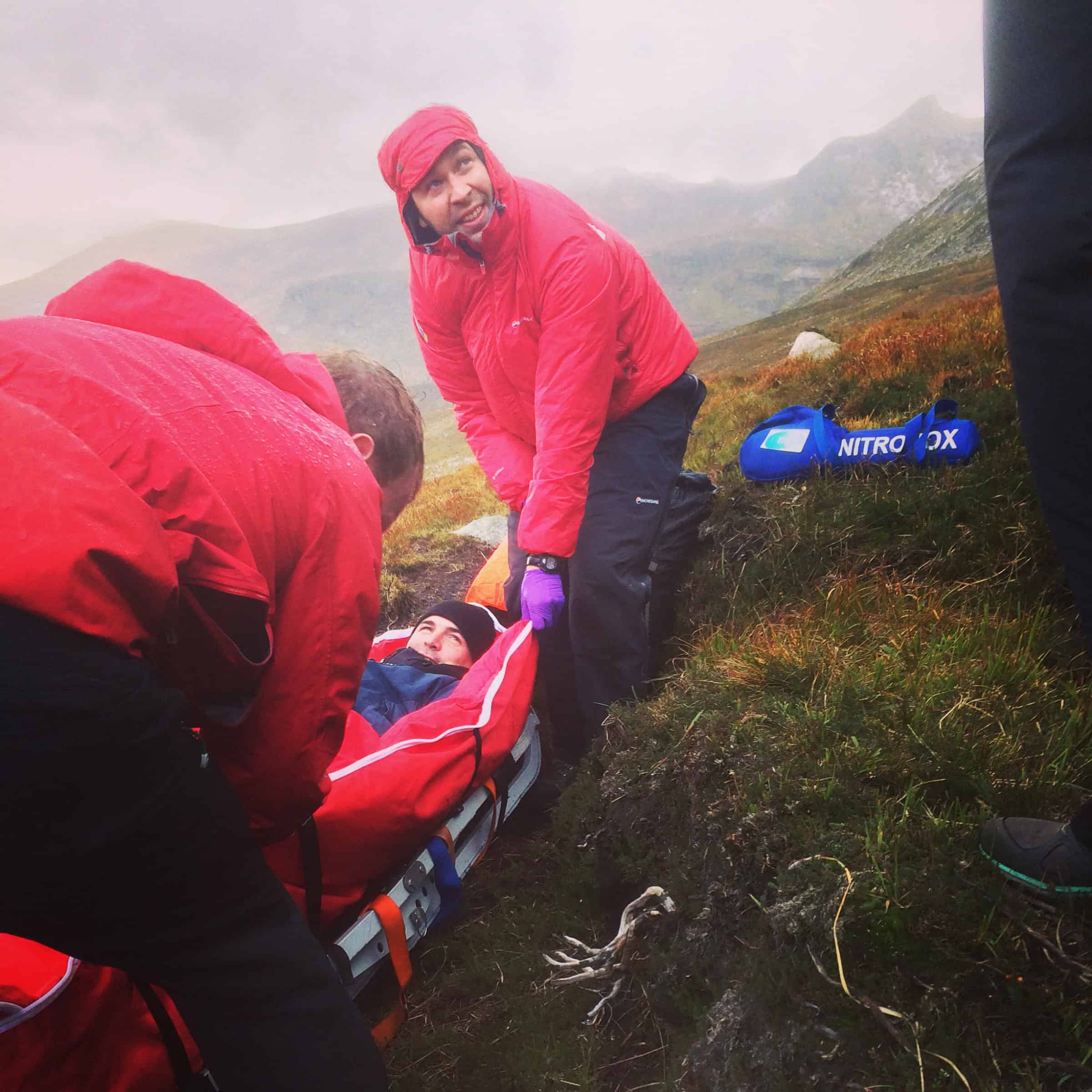 Two camps and a rescue: A wild weekend in the Mournes Rescue team carrying a hiker on a mountain trail in rainy weather, outdoor emergency rescue, rugged landscape, adrenaline-filled mountain rescue scenario.