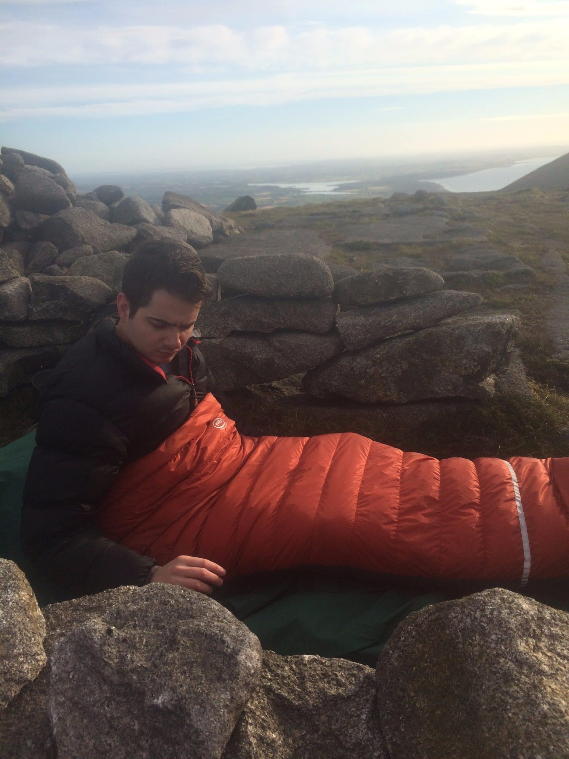 PHD Launches Sleep Systems Protected outdoor camping scene in the mountains with a hiker wrapped in an orange sleeping bag, rocky landscape, scenic view of lakes, hills, and open sky, ideal for hiking, trekking, and outdoor adventure enthusiasts.