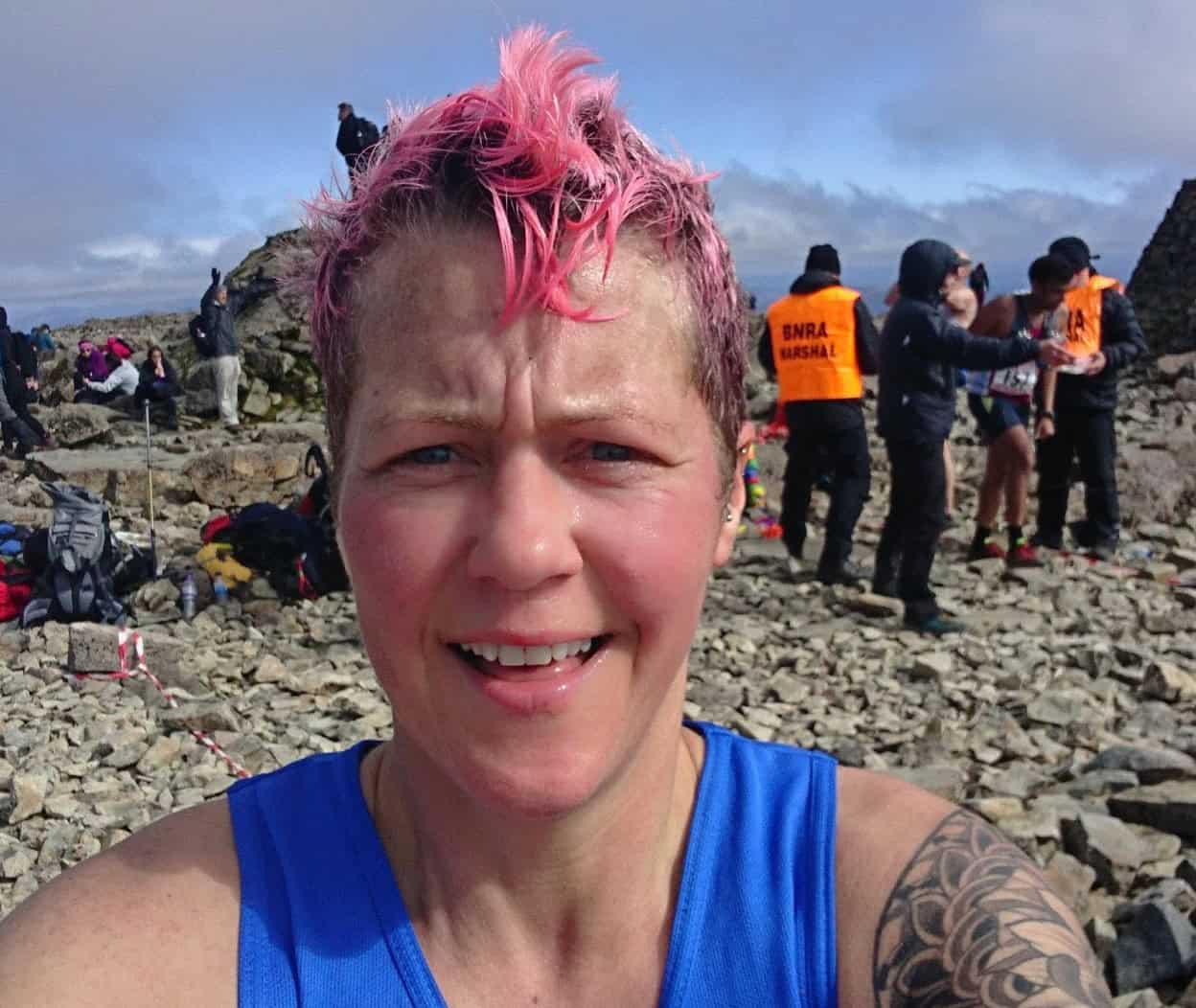 The Ben Vibrant female trail runner with pink hair smiling after a mountain race on rocky terrain at high altitude. Participants and marshals are visible in the background under cloudy skies.