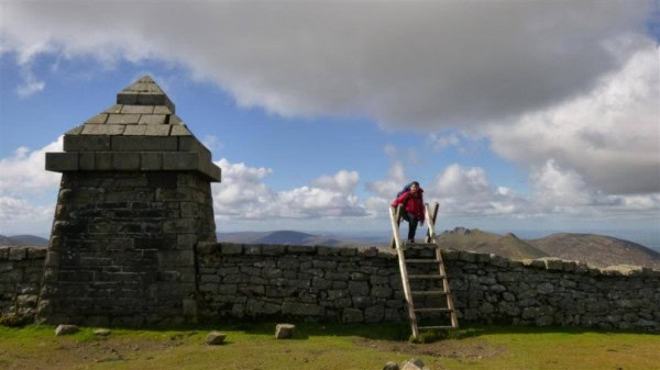 Two nights of the Mournes at Commedagh and Tollymore A hiker climbing a wooden ladder over ancient stone ruins during a scenic outdoor adventure under a partly cloudy sky.