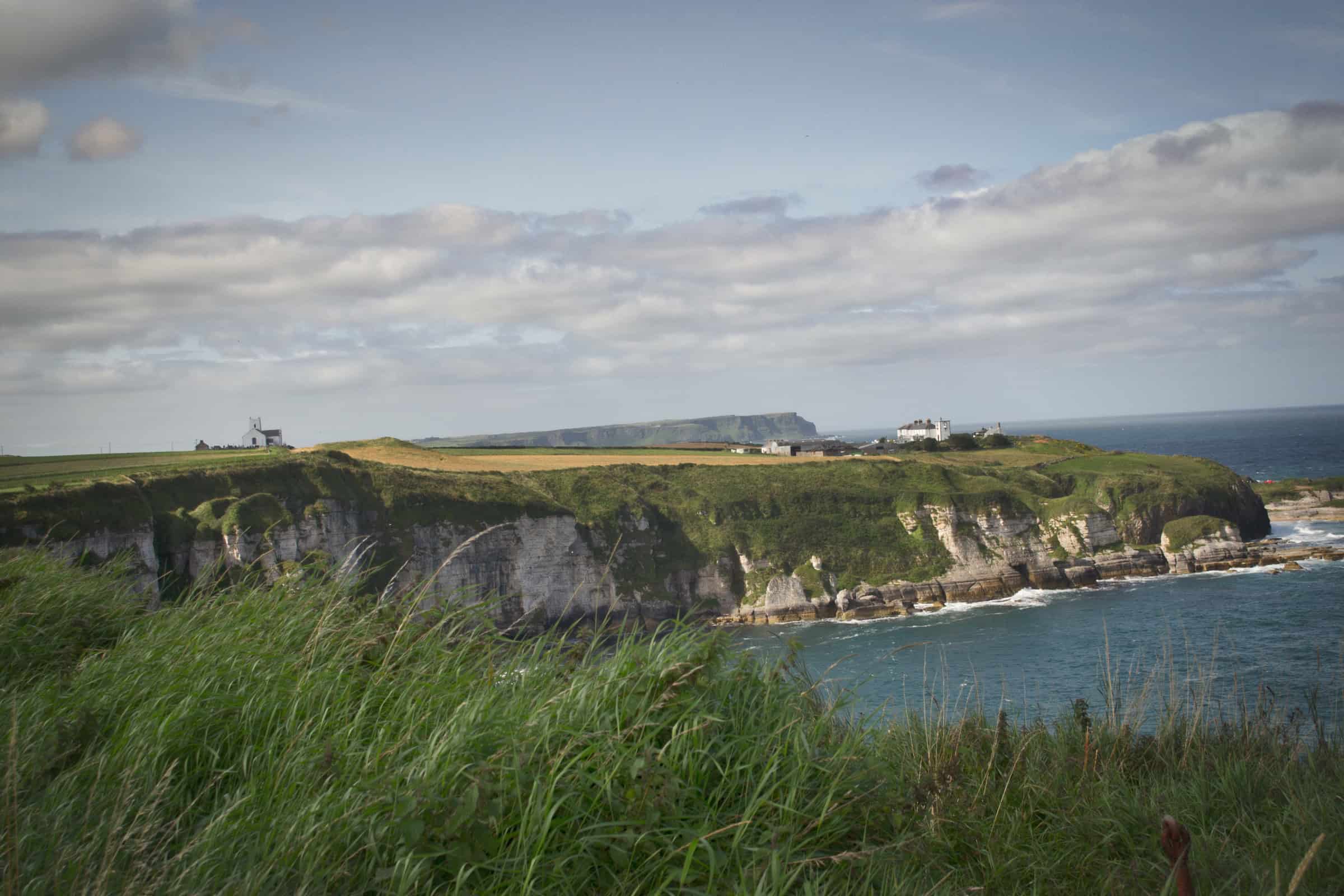 The Causeway Coast Way Vast coastal cliffs with green grass and white limestone formations overlooking the Atlantic Ocean on a cloudy day.