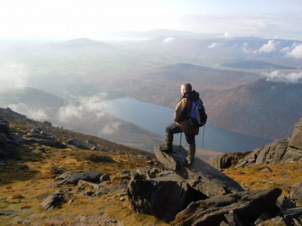 Photo Album: John’s Mourne Photo Collection Hiker standing on rocky mountain ledge overlooking a tranquil lake and expansive valley in the distance during a scenic outdoor adventure.