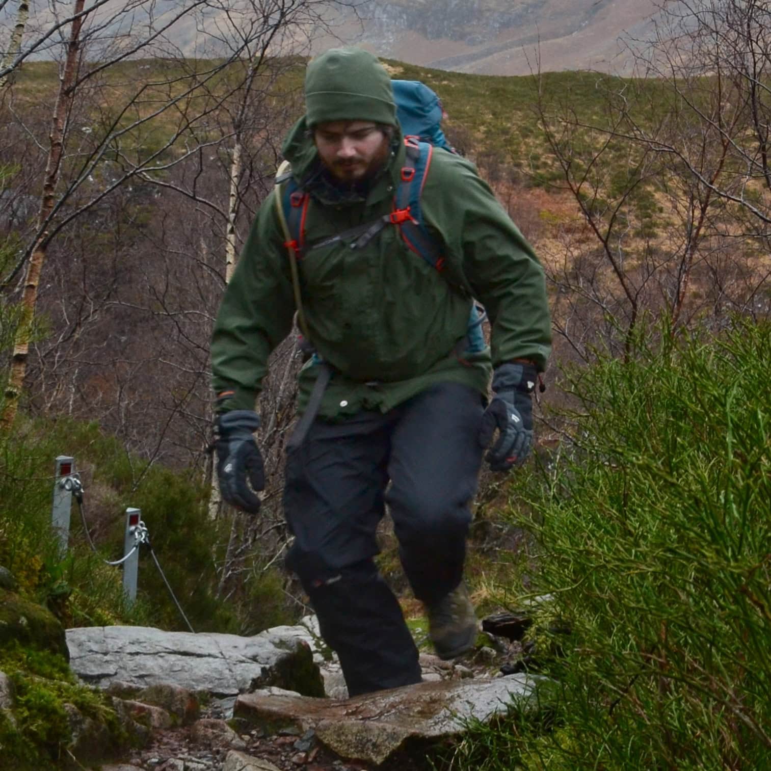 Beginner’s Guide to Layering Clothing Hiking enthusiast crossings rocky trail in scenic mountain landscape with trees and shrubs, wearing outdoor gear and backpack, on a cloudy day.
