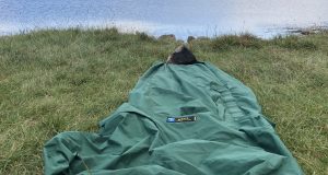 Tips For Bivvy Bag Newbies A green sleeping bag laid out on grassy terrain beside a calm body of water, ideal for outdoor camping and hiking adventures. Perfect for nature lovers seeking tips on wilderness accommodations.