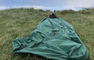 Tips For Bivvy Bag Newbies A green sleeping bag laid out on grassy terrain beside a calm body of water, ideal for outdoor camping and hiking adventures. Perfect for nature lovers seeking tips on wilderness accommodations.