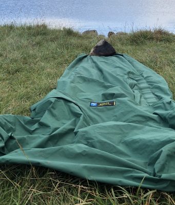 Tips For Bivvy Bag Newbies A green sleeping bag laid out on grassy terrain beside a calm body of water, ideal for outdoor camping and hiking adventures. Perfect for nature lovers seeking tips on wilderness accommodations.