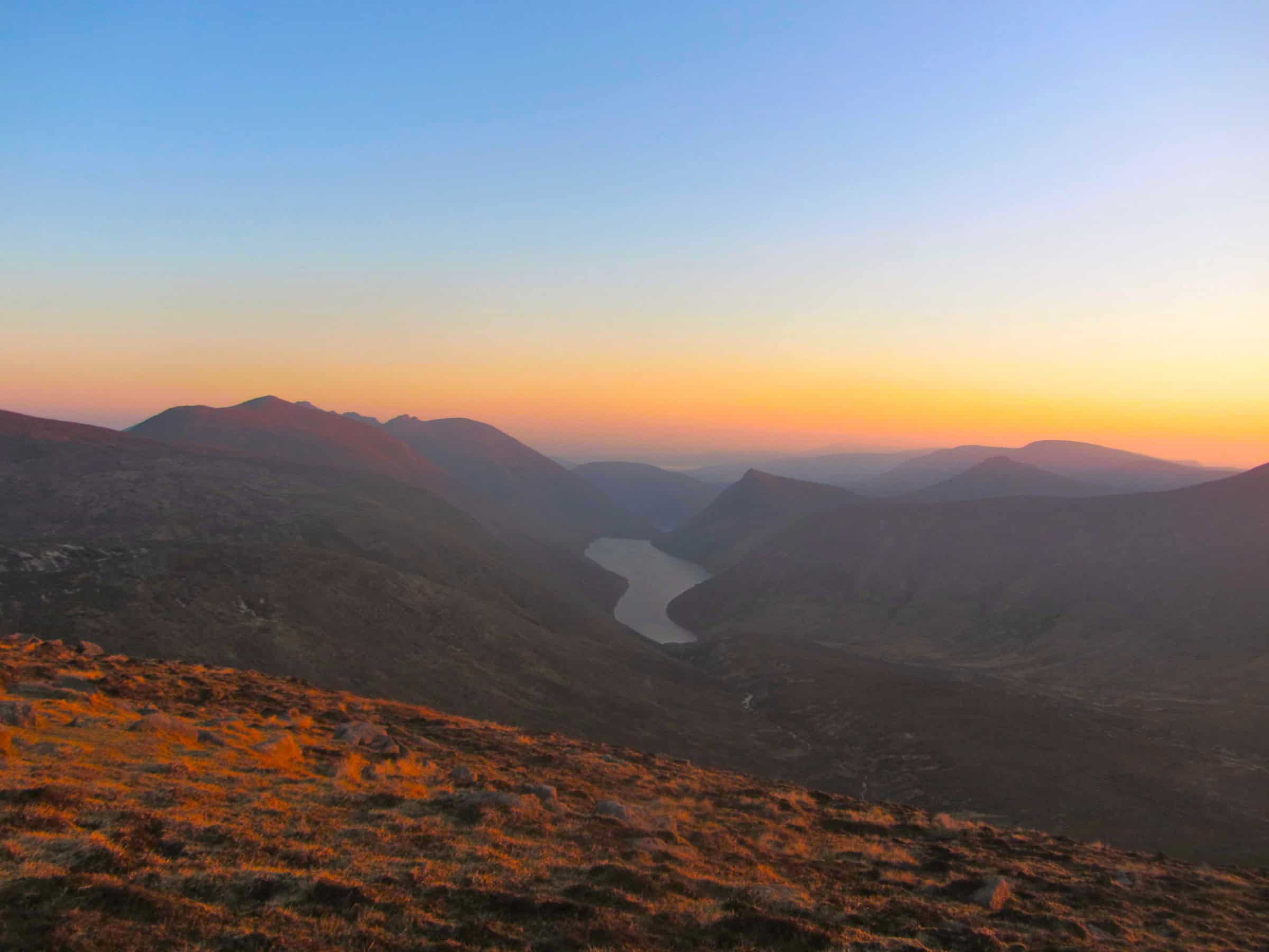 Photo Album: Slieve Corragh Summit Camp Breathtaking mountain landscape during sunset with a winding river in a remote wilderness area, ideal for hiking and outdoor adventures.