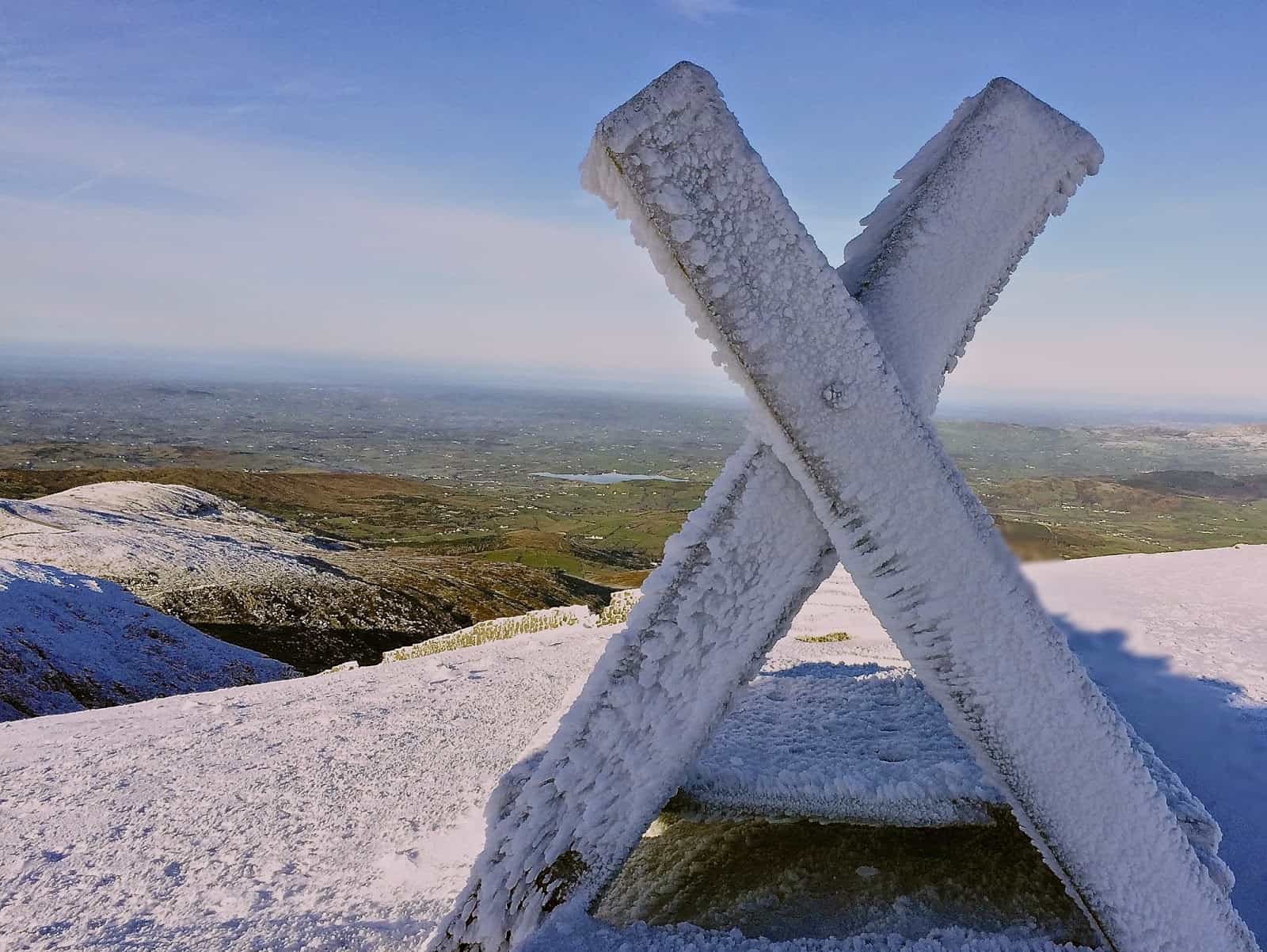Snow in the Mournes Frost-covered mountain summit marker with a scenic view of valleys and hills in the background, under a clear blue sky, emphasizing winter hiking adventures.