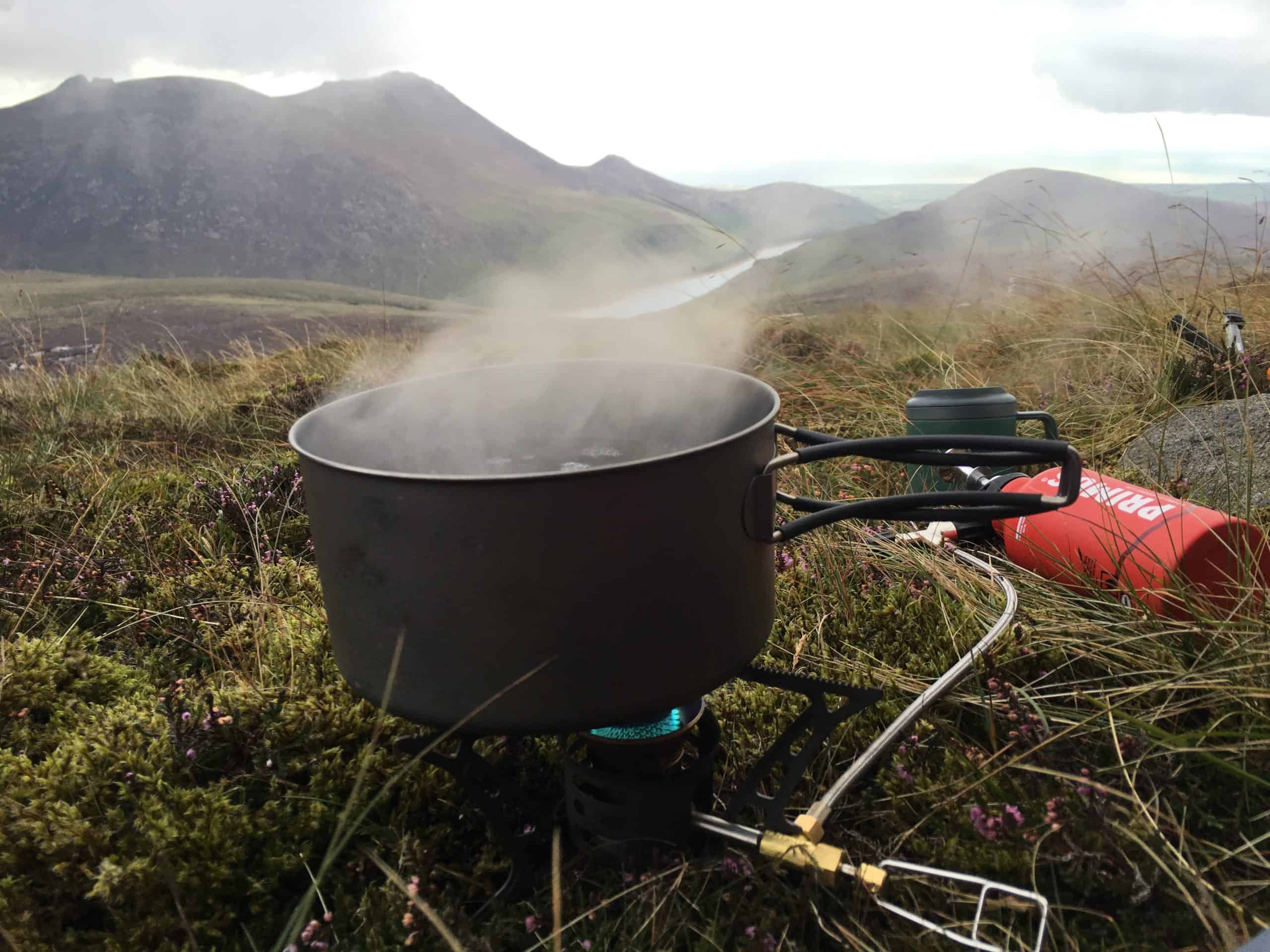 Primus OmniLite Ti Review A compact camping stove with a boiling pot on a mountain hillside, surrounded by grass and wildflowers, with misty mountains and cloudy sky in the background.
