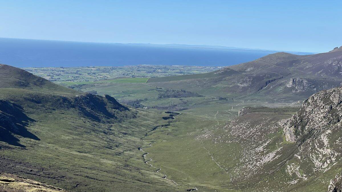 Vast mountainous landscape with rolling green hills, rugged rocks, and a distant view of the ocean on a clear, sunny day. Ideal for hiking and outdoor adventure enthusiasts exploring nature trails and scenic routes.