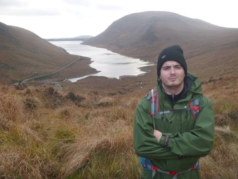 Silent Valley Circuit via Carn and Muck Serious male hiker in green jacket with backpack standing in scenic mountain landscape with lake and rolling hills in the background.