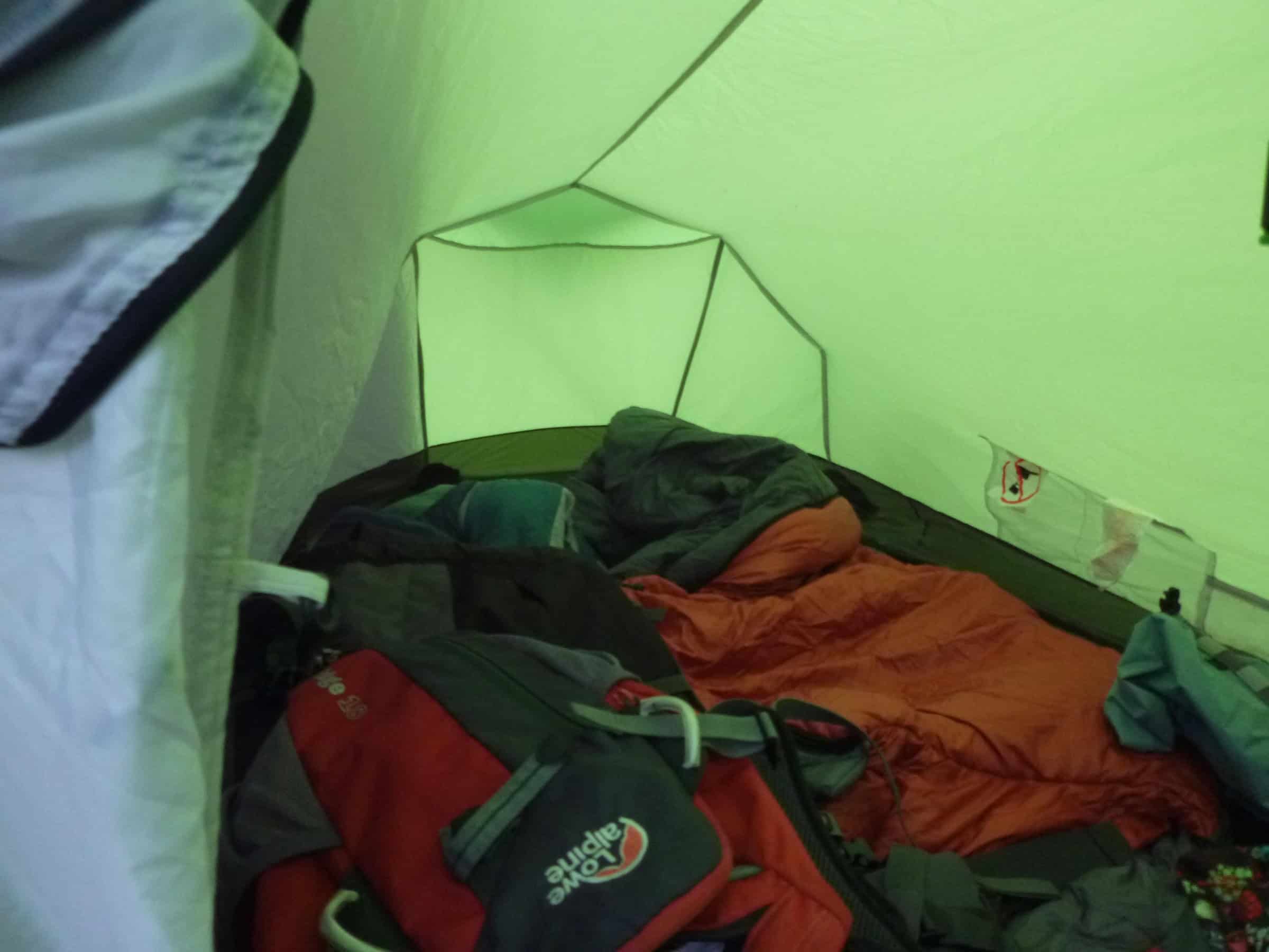 A young hiker setting up camping gear on a grassy hillside overlooking the ocean on a cloudy day, with a bright green Coleman tent nearby, embodying outdoor adventure and scenic backpacking experiences.