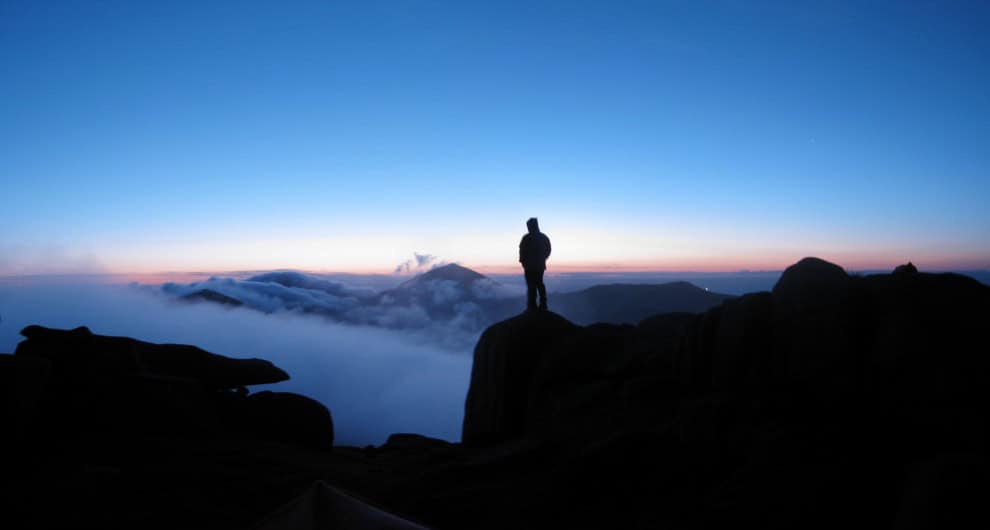 Binnian summit camp Silhouette of a hiker standing on a rocky mountain summit during sunrise with scenic mountain views and clouds beneath, perfect for outdoor adventure and hiking enthusiasts.