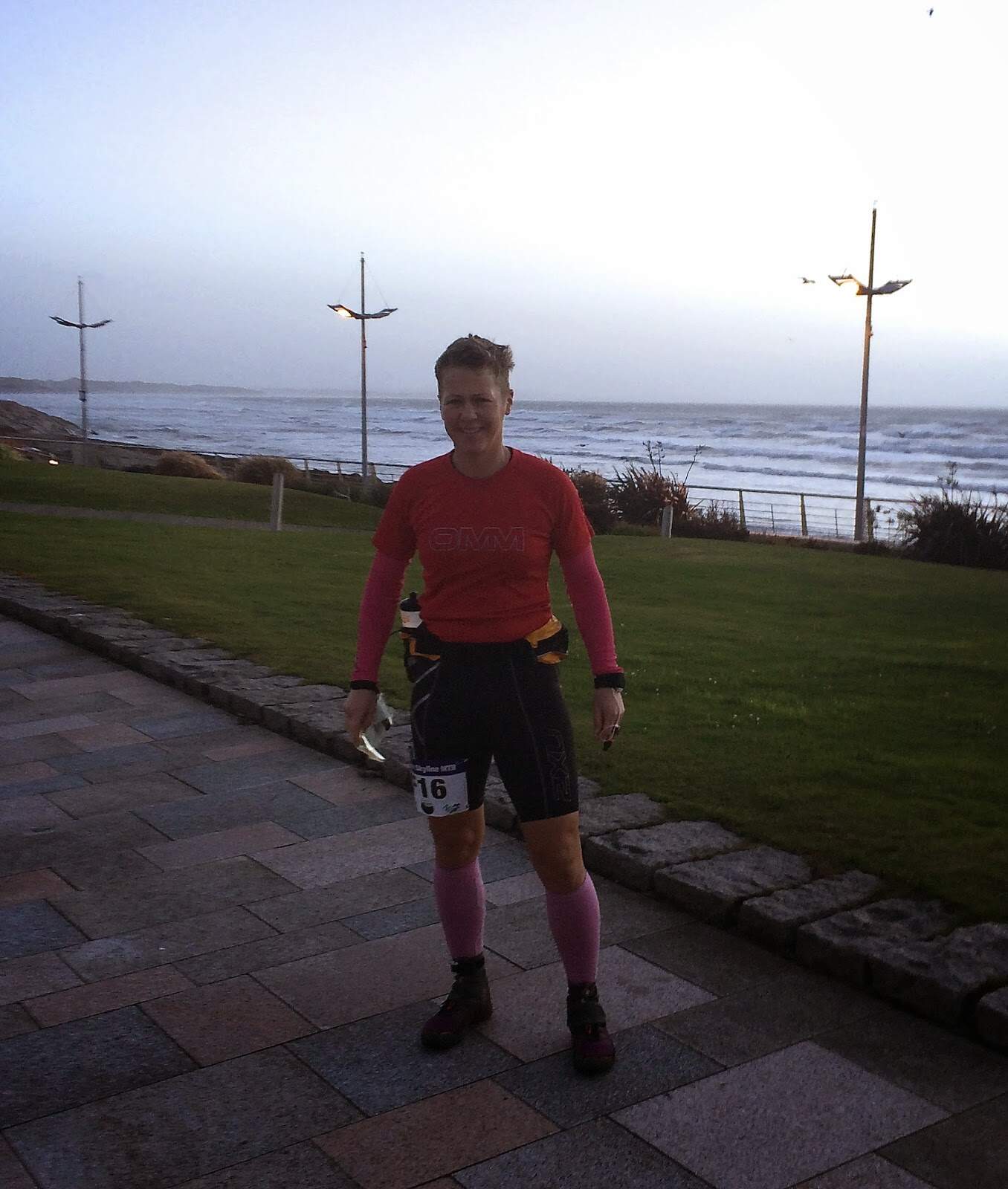 Mourne Skyline MTR Race A woman in athletic gear standing outdoors by the seaside with wind turbines in the background, ready for trail running or outdoor adventure.