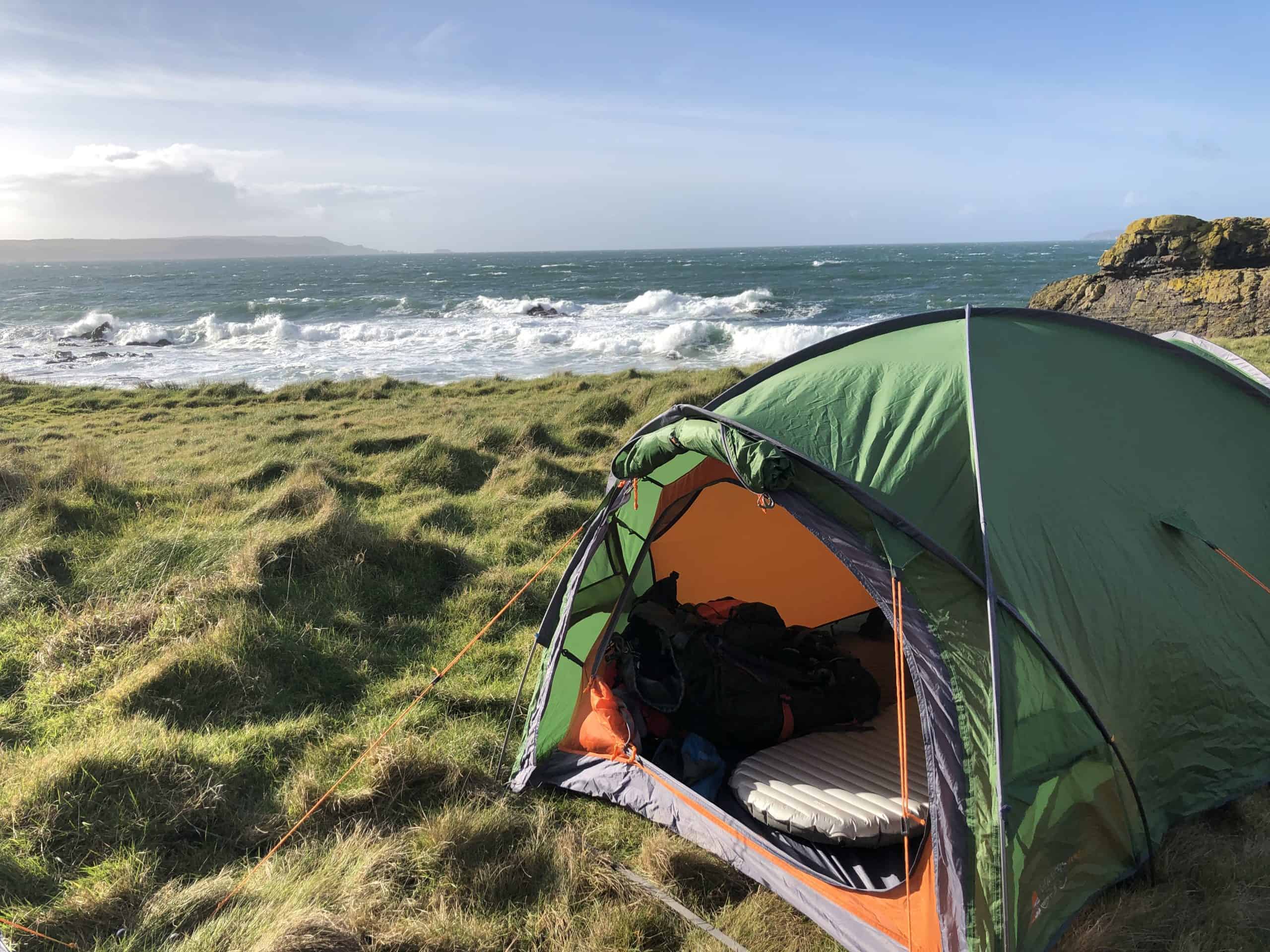 Ballycastle Wild Camp Quiet outdoor camping scene with a green tent on grassy coastal land, overlooking the ocean with waves, rocks, and a partly cloudy sky. Perfect for hiking, adventure travel, and nature excursions.